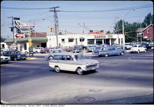 Car dealership showing low white building and a parking lot decorated with lots of pennants.