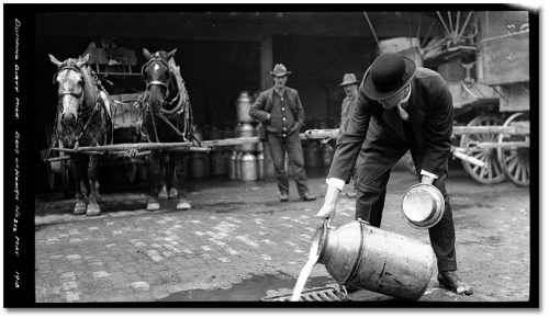 Man in suit and hat pours milk from large tin into sewer grate.