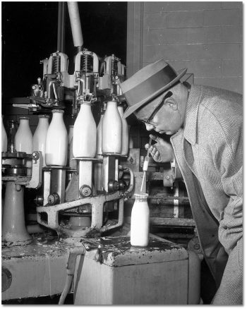 A man uses a syringe to pull a sample of milk out of a bottle, with an assembly line of bottles in the background.