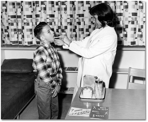 In a medical office, a woman in a lab coat opens a boy's mouth with a tongue depressor.