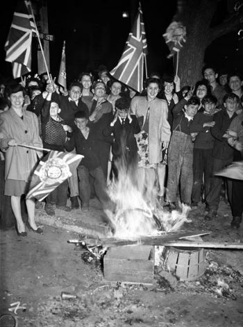 Adults and children wave flags and watch a small bonfire.