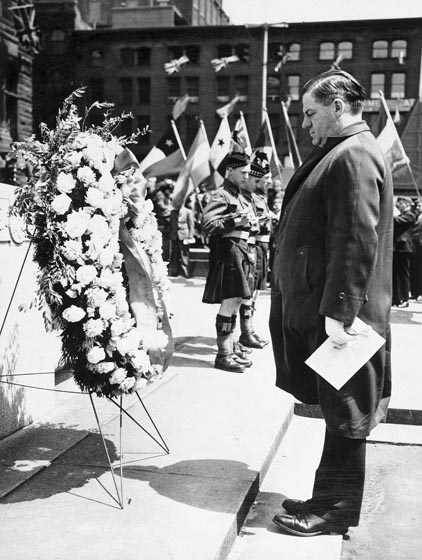 Man in dark coat stands in front of wreath made of flowers..