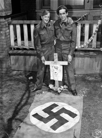 On a lawn, two men in army uniforms display a mock grave with a Nazi flag and a cross gravemarker that reads To Hell With Hitler.