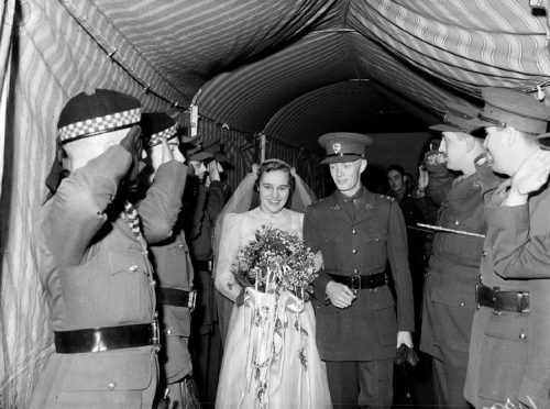 A bride and a soldier walk between two rows of people under a tent.