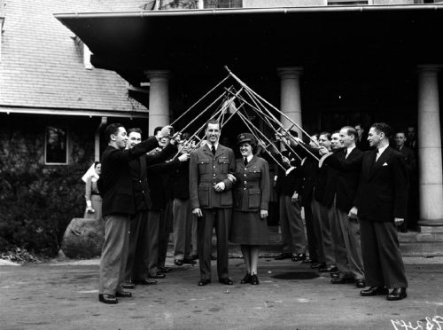 A man and woman in armed forces uniforms walk under an arch of crutches and canes held up by soldiers.