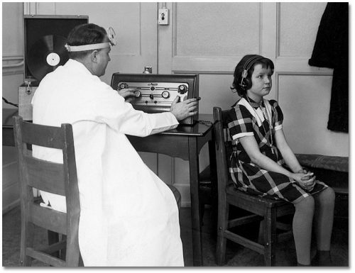 A girl with earphones on sits in a chair while a man at a desk beside her operates dials.