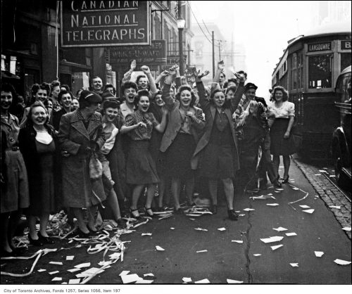 A large group of women is standing on the sidewalk, cheering and waving.