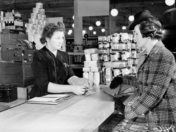 A customer purchases something at a counter stacked with bags of coffee. The cashier is tearing a ticket out of a book of ration stamps.