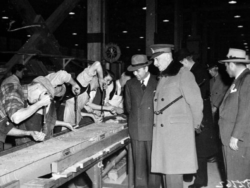 Workers work at a long bench while men in suits look on.