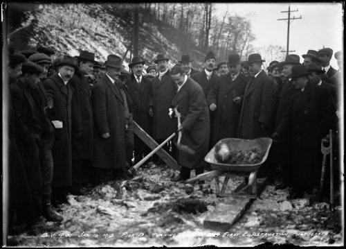 Men in suits and coats surrounding a man shovelling earth into a wheelbarrow.