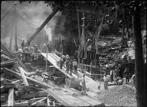 Busy construction site with men transporting supplies up a wooden ramp in wheelbarrows.