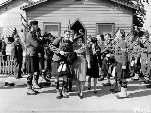 A woman in a dress and a man in a kilt walk between two rows of soldiers with bagpipes.