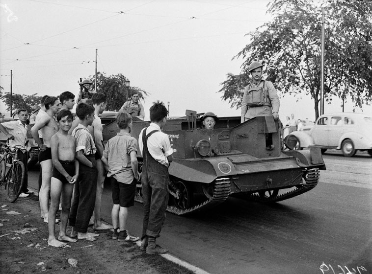 Boys in casual clothes and bathing suits watch as soldiers drive along the road in a tank-like gun carrier.