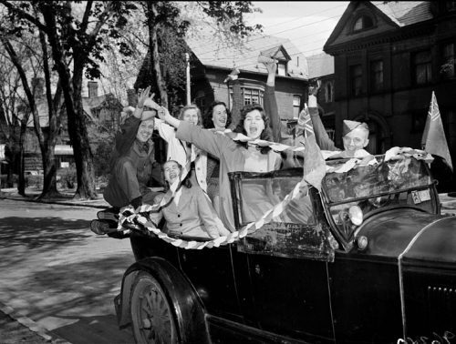 University students cheer in a car decorated with flags and streamers.