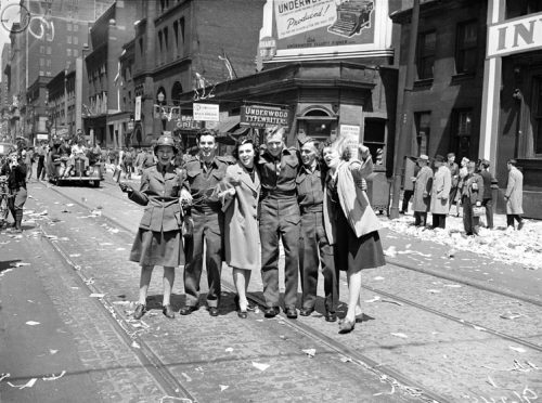 Six men and women, some in uniform, stand in a row, smiling and cheering at the camera. Some are waving flags. The street is covered in tickertape and other paper.