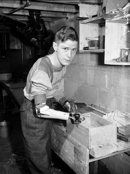 A young man with an artificial right arm and hand is planing a wooden box smooth.