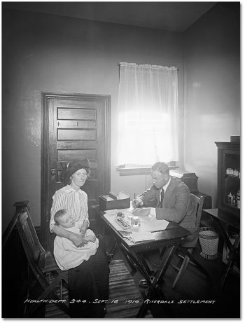 Sitting in front of a desk. a woman holds a baby on her lap, while the doctor on the other side of the desk looks at the baby and writes something on a piece of paper.