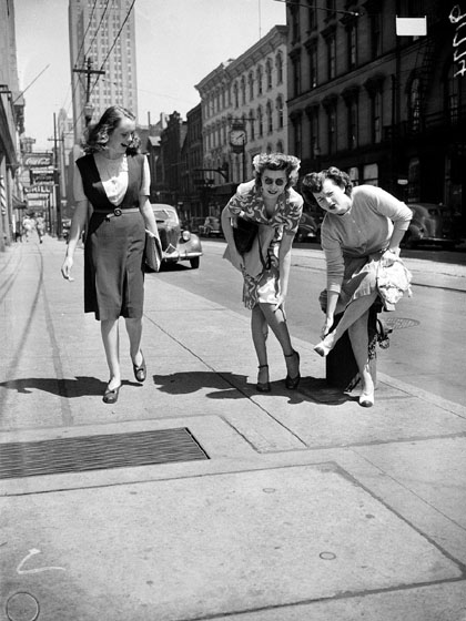 Two women standing on a sidewalk bend over to hold their feet and legs, and make faces to show how much their feet hurt. Another woman laughs as she walks past them.