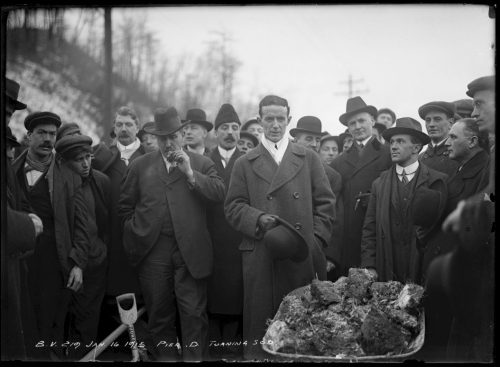 Men in suits and coats surrounding a wheelbarrow full of clods of earth.