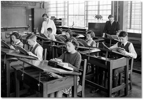 Children read books in a classroom with large windows and slanting desktops. Some are wearing glasses.