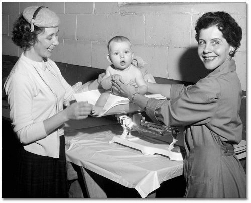 A mother and a nurse smile and hold a baby on a scale.