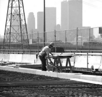 Standing on deck of expressway, worker uses circular saw to cut something on worktable