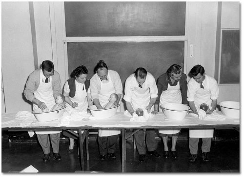 Men wearing apron bathe baby dolls in basins on a table.