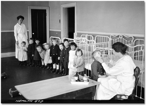 Nurse looking into a child's mouth while a line of children looks on.