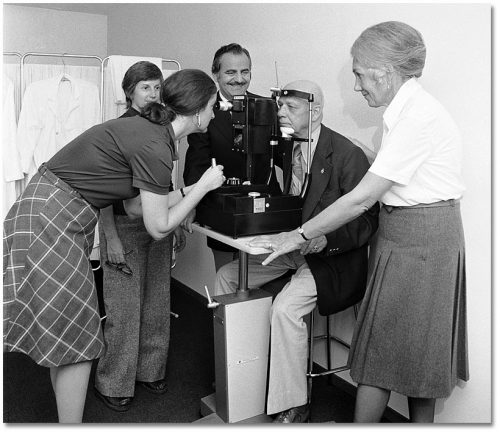 An optometrist examines an elderly man's eyes using a machine while other people watch.