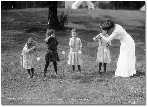 Four girls stand on the grass with mugs in their hands, brushing their teeth, while a teacher helps one of them.