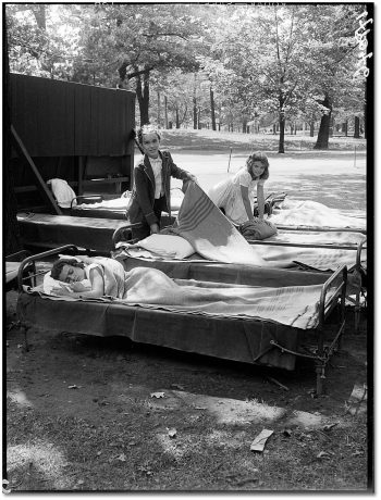 Girls fold blankets over cots outdoors under some trees, while a girl sleeps in a third cot.