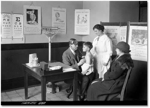 A doctor listens to a boy's chest with a stethoscope in a corner of a room with illustrated posters about healthy habits all over the walls.