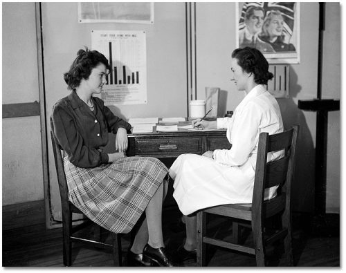 A nurse in a white lab coat and a teenage girl wearing a plaid skirt talk at a desk while the nurse takes notes.