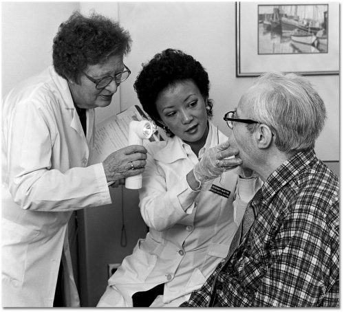 Two dentists in white lab coats examine an elderly man's teeth.