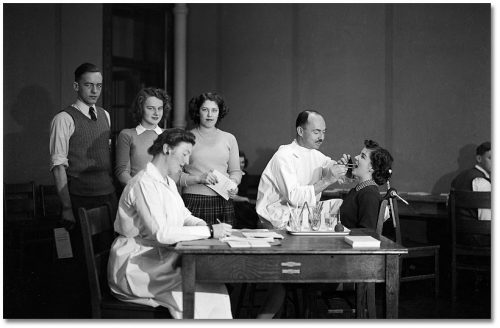 A dentist looks in a teenage girl's mouth, while a line of teenagers stands beside a woman in a lab coat taking notes at a desk.