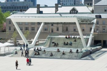 View of the theatre stage in Nathan Phillips Square, where people can be seen sitting on the steps eating their lunch and enjoying the sunny weather