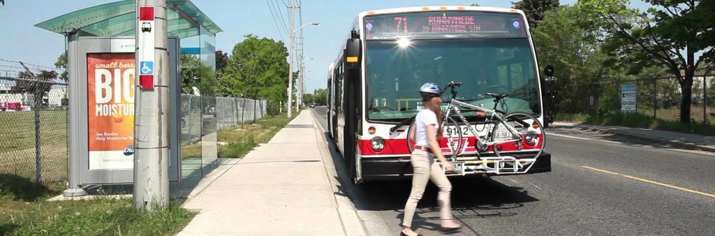 Image of a stopped TTC Bus. A women is hooking up her bike to the front of the bus.