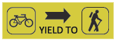 A wide yellow rectangle with a black bicycle on the left most side, an arrow pointing to the right in the middle with the words Yield To underneath, and a black stick figure walking with a stick on the right. 