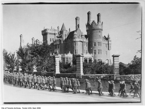 A column of soldiers walk past a large building with towers and turrets.