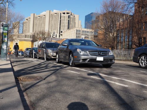 A person cycles in a cycle track on Hoskin Ave that is separated from motor vehicle traffic by parked cars and bollards. 