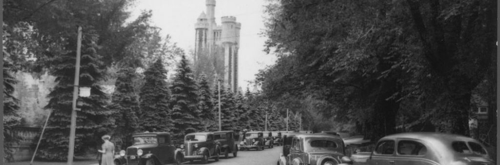 A black and white photograph showing a tall, castle-like tower visible through an opening in the trees.