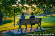 In Riverdale Park, two people sitting on a bench taking in the view of the sunlight.