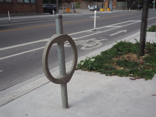 A post and ring bicycle rack located on the sidewalk beside a bicycle lane