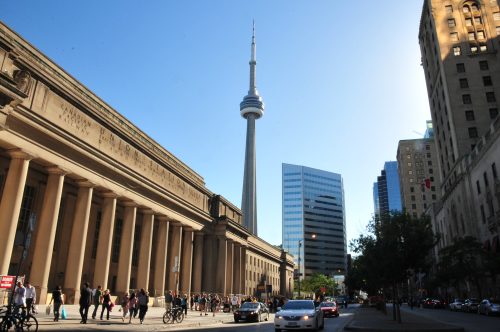 Front of the Union Station building with the CN Tower in the background and pedestrians walking and cycling in front of the building. Photo taken from Bay and Front Street.