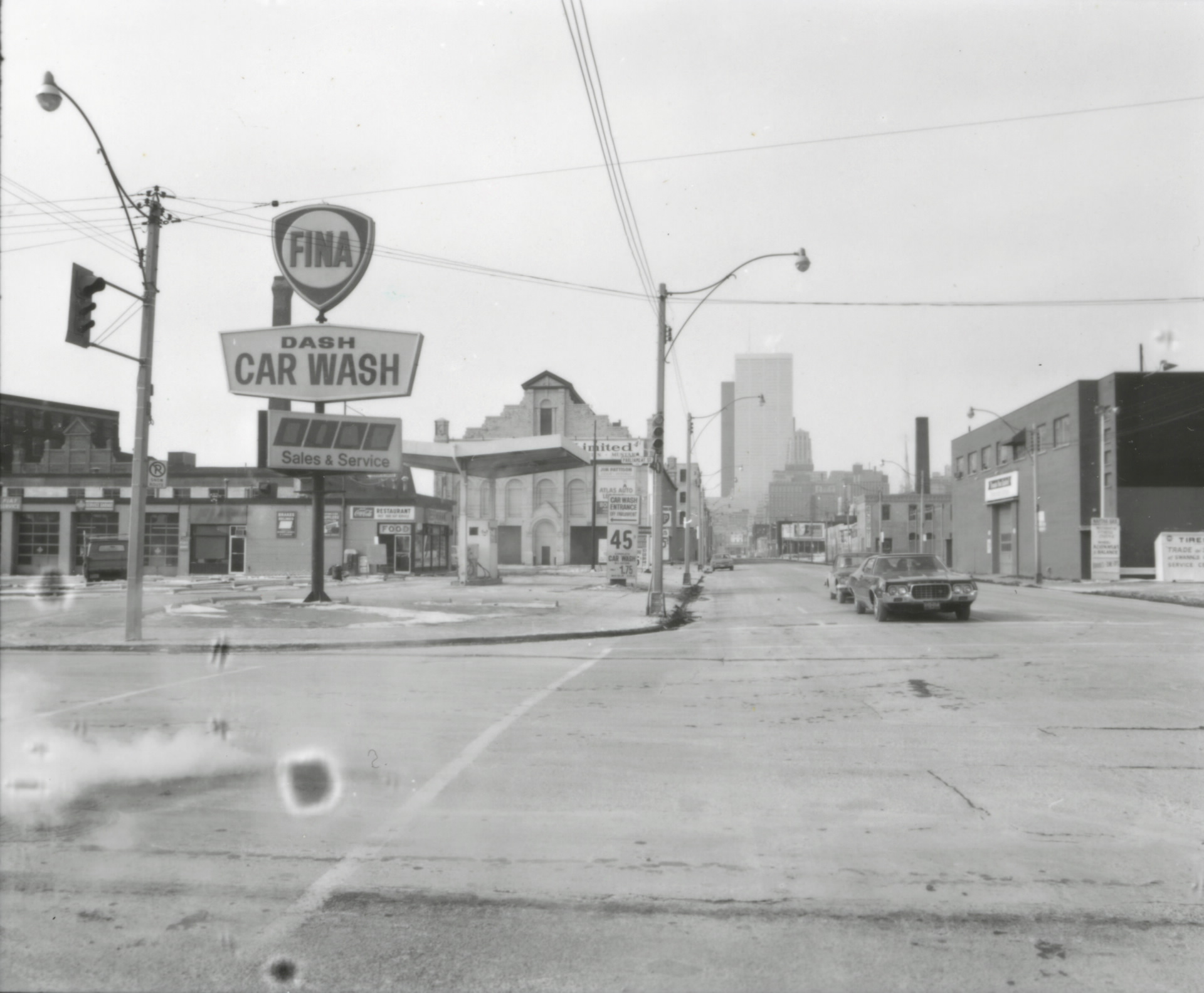 View of Front Street, looking west from Parliament Street, with a car wash and gas station in view on the left, cars on the street waiting at a light and downtown can be seen in the horizon.