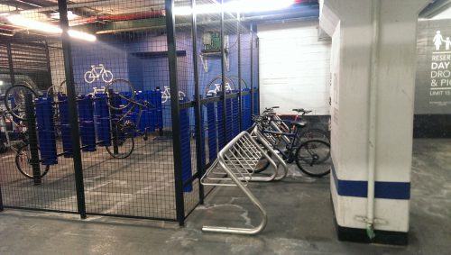 Long term bicycle parking located in the garage of an office building. A secure bicycle cage is located beside some bicycle racks.