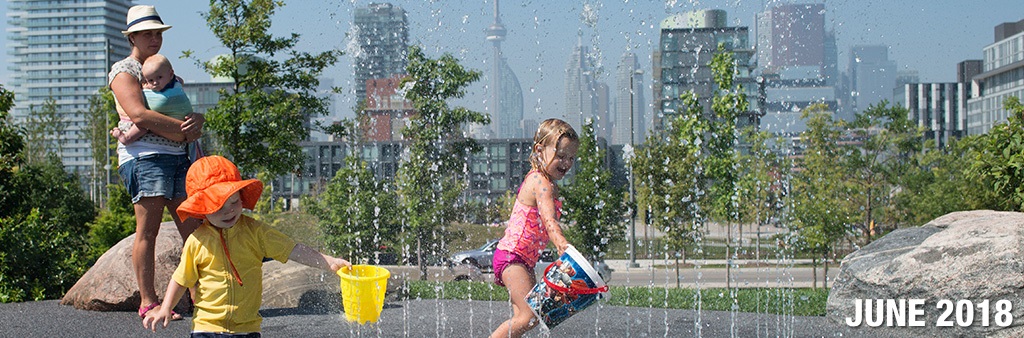 A young girl and boy play in a City splash pad as their mother watches holding their baby brother. The City skyline is in the background.