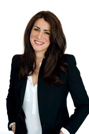 Photograph of Magali Simard standing and smiling wearing a black suit with a white shirt. Her hands are in her pockets.
