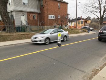 Image of a residential street with 1 lane of traffic going each way. On the painted yellow centre line, there is a in road flexible speed sign that's 3 feet high and reads maximum of 40 km/h. 