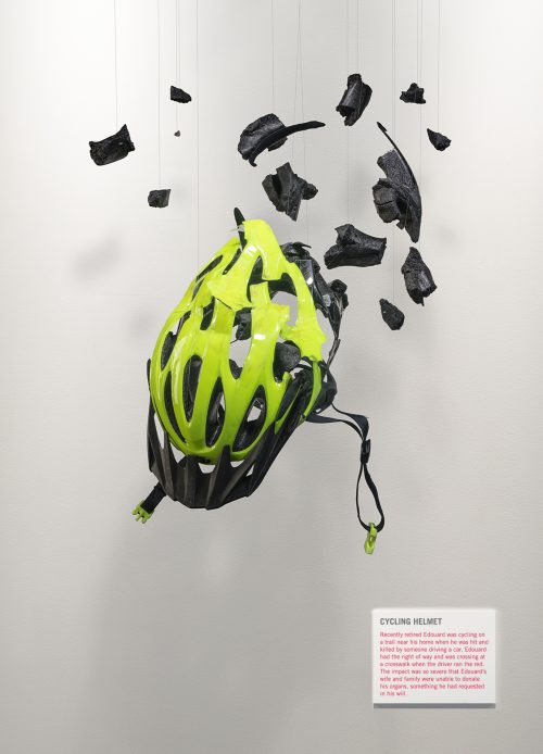 A damaged cycling helmet is suspended in mid-air like an exhibit in an art gallery. Small fragments hang around the helmet appearing to be frozen in mid-air.